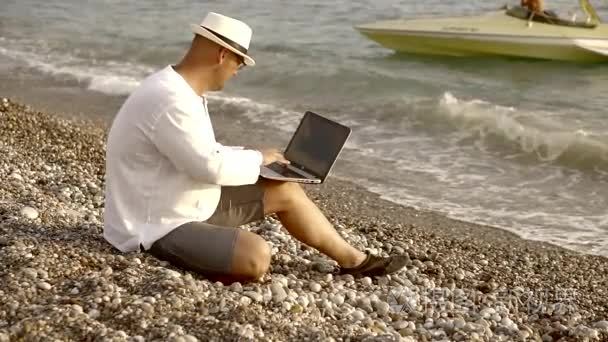 成年男性穿着白色衬衣, 戴着眼镜, 在日落时坐在海边的电脑上工作。他喜欢自然和放松。视频