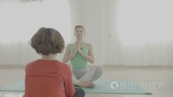 教瑜伽的年轻美丽的妇女给一个小女孩和如何打坐在健身室的垫子上