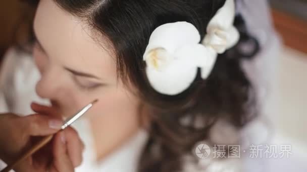 顶部视图。专业化妆艺术家正在应用婚礼化妆在美丽的黑发新娘的脸上与兰花在她的头发