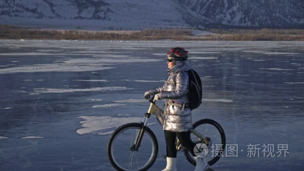 女人在冰上骑自行车。这个女孩穿着一件银色羽绒服  骑自行车背包和头盔。冰冻的贝加尔湖的冰。自行车上的轮胎布满了特殊的尖峰。