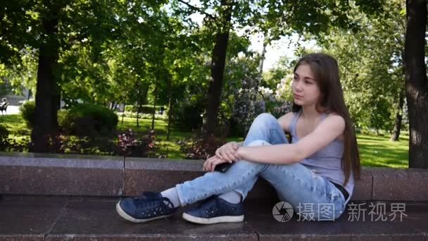可爱的十几岁的女孩检查电话和等待的电话, 坐在公园。视频素材高清拍摄静态相机