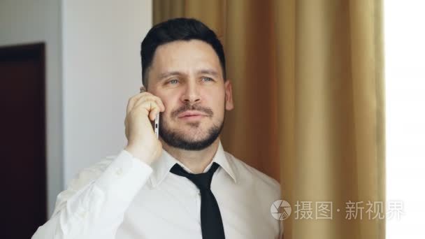 成人微笑的商人在白色衬衣和领带谈话在移动电话站立在窗口附近在旅馆房间里