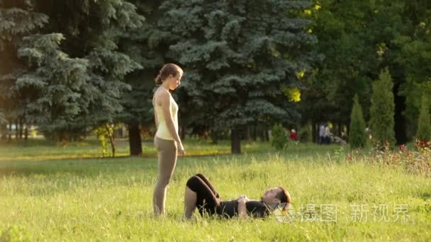 美丽的情侣练习瑜珈。年轻的瑜伽导师在绿色草地上的城市公园练习。两个成功的年轻人进行瑜珈练习。男人和女人在日落时学习瑜伽