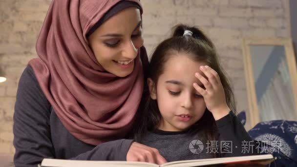 戴头巾的年轻女子和女儿坐在沙发上, 教她如何阅读, 读书, 幸福的家庭概念, 关闭 50 fps视频