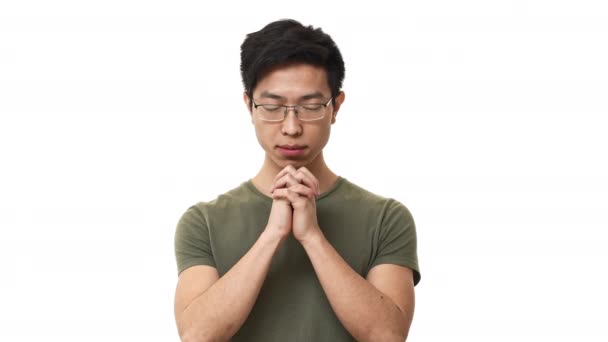 年轻的亚洲男子戴眼镜和基本 t恤的肖像, 双手合十, 闭着眼睛祈祷, 在