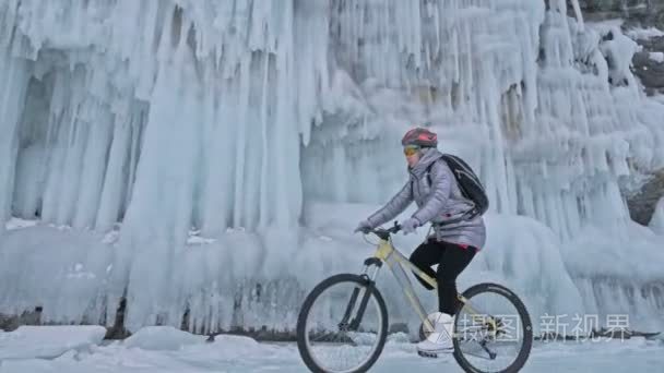 女人在冰窟附近骑自行车。岩石与冰洞穴和冰柱是非常美丽的。这个女孩穿着银色羽绒服  骑自行车背包和头盔。旅行者是骑自行车
