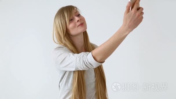 现代青年。美丽的长头发的欧洲前瞻的女孩与金发自拍在智能手机附近的白色墙壁