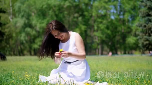 夏天  美丽的黑发女人穿着白色的连衣裙  坐在绿色的草坪上  小草  编织着黄色蒲公英的花环  微笑着