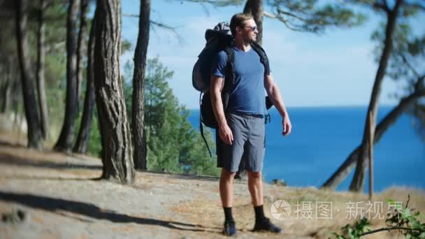 一个英俊的旅游男子与一个大背包旅行到陡峭险峻的海岸边与松树林的边缘。以壮丽的海景景观。4k