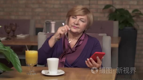 一个迷人的中年妇女坐在咖啡馆的桌子旁  拿着电话。她戴上眼镜  开始滚动手机