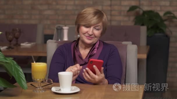 一个名叫一个漂亮的中年妇女坐在咖啡馆里看着电话笑着的特写镜头。桌子上有满满一杯橙汁和一杯茶