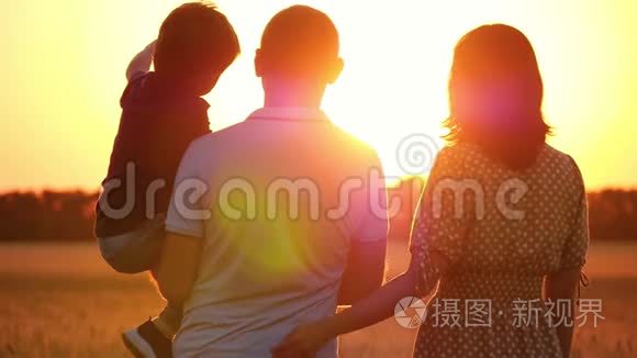 幸福的一家人看着夕阳，站在麦田里。 抱着孩子的男人.. 女人拥抱男人