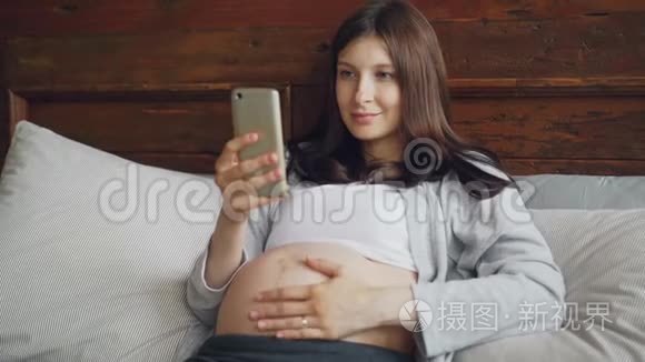 美丽的怀孕少女黑发正在抚摸她的婴儿肿块，并使用智能手机在家里的床上轻松愉快的微笑
