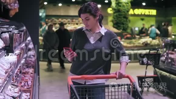 年轻的深色头发的女顾客在杂货店里穿过肉道时看着手机。 女孩带着腊肠