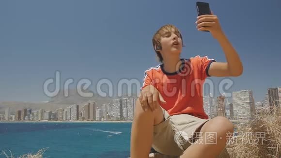 可爱的青少年男孩在智能手机上打视频电话，展示海上城市天际线背景。