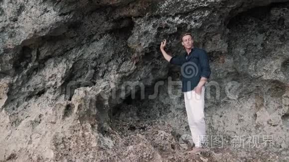 穿着白色裤子和黑色衬衫的白人男子站在靠近岩石的海滩上，向远处望去