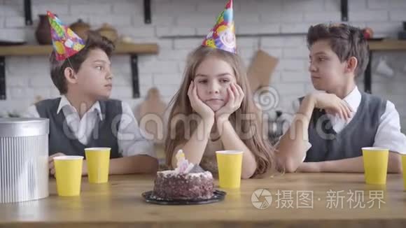 一幅美丽的小悲伤女孩坐在桌子旁，双胞胎兄弟戴着派对帽的肖像。 高加索少年