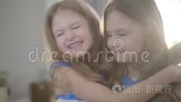 幸福的小漂亮女孩的特写脸拥抱双胞胎妹妹，亲吻她的脸颊。 两个孩子在阳光下摆姿势
