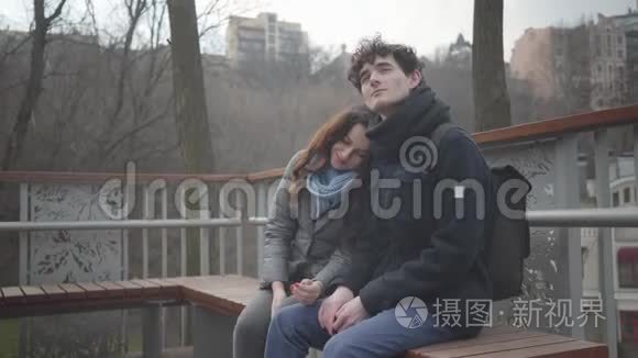 快乐的白人夫妇坐在公园的长凳上微笑。 女孩抱着头`男孩的肩膀和男人看着