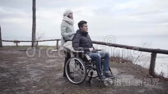 年轻积极的白人妇女触摸轮椅上残疾男子的肩膀并指向别处的肖像。 强国