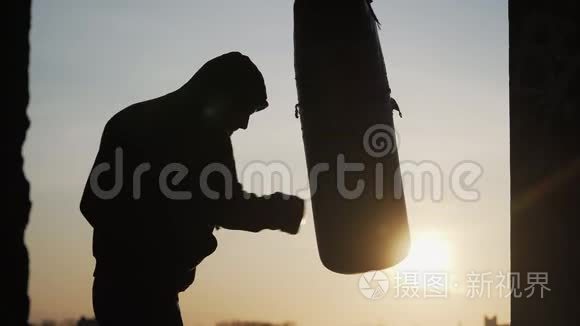 剪影男童拳手在美丽的夕阳背景下跳动着一个拳击袋。 体育和武术。 户外拳击训练