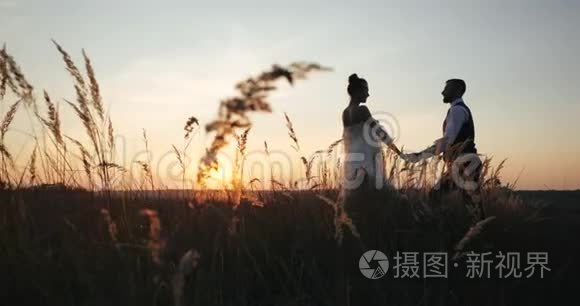 时尚的千禧新娘和新郎在傍晚的秋天，迎着夕阳，沿着小穗田野走着。 欧洲