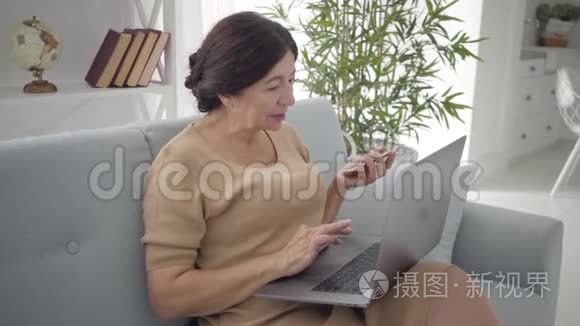 自信的高加索族妇女使用笔记本电脑和智能手机坐在室内沙发上。 成功的黑发中