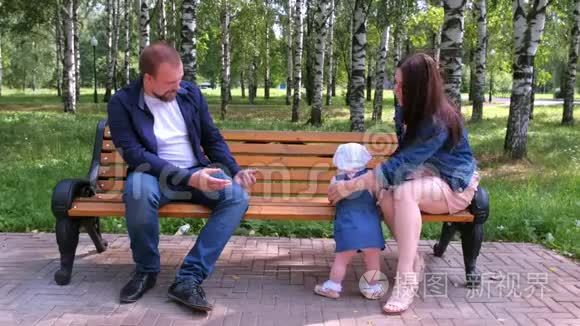 小女孩试图从妈妈到爸爸坐在公园的长凳上迈出第一步。