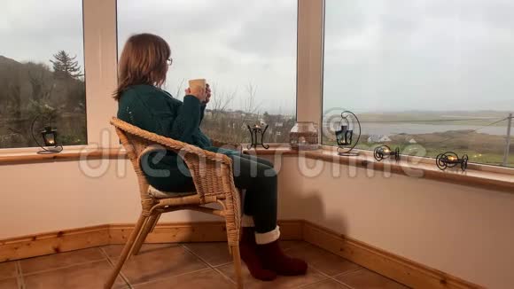 一个孤独的女人坐在阳台上的一扇宽窗旁舒适的柳条椅子上,喝茶,享受