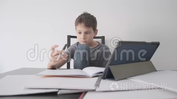 悲伤的孩子坐在桌子上，拿着平板电脑和许多书。 男孩感到无聊，因为他不懂作业