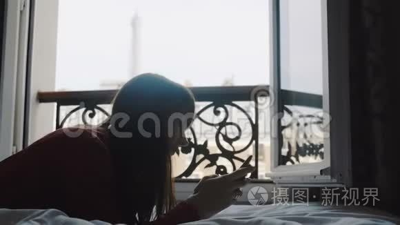 快乐美丽的欧洲女人在智能手机应用程序上输入信息，躺在阳台床上，埃菲尔铁塔观景慢