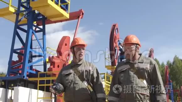 两名男性石油工人在油泵千斤顶附近行走和交谈。 石油工程师监督原油钻井平台