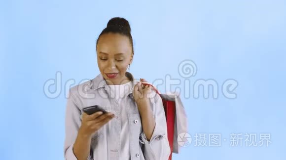 美国黑人妇女带着购物袋的肖像正在蓝色背景下通过智能手机录制语音信息。