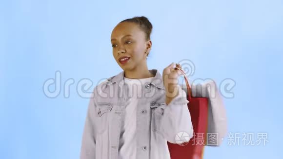 美国黑人妇女带着购物袋的肖像正在工作室的蓝色背景上通过智能手机交谈。 珍妮夹克