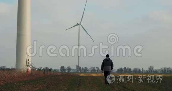 一个男人在后台旋转风车的田野里行走。 风的力量。 清洁和可再生能源解决方案。 风力涡轮机