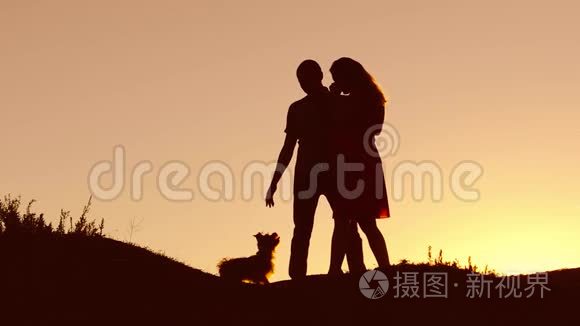 恋爱中的情侣在自然日落和接吻时跳舞剪影。 爱的男人和女人与狗跳舞剪影缓慢