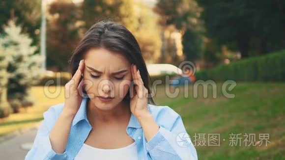 一个穿着蓝色短衫的年轻生病的女人在城市公园的背景下抚摸着她的头。 快关门。 复制空间