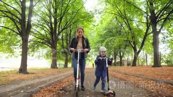 4K镜头，快乐的微笑男孩和年轻的母亲骑着滑板车在美丽的小巷与高树在公园