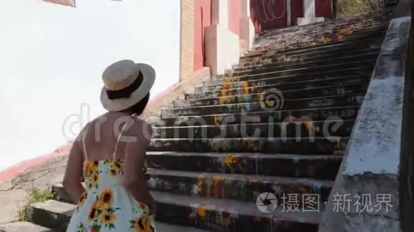 年轻女子戴着草帽，穿着夏装，带着带子爬上城市里粉刷过的楼梯。 关闭后方视野