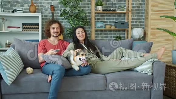 年轻的家庭和科吉狗在家沙发上看电视，笑着放松