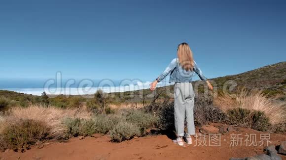 穿着牛仔夹克和条纹裤子的金发女人站在云层上方的一座高山上，举起双手。 这就是