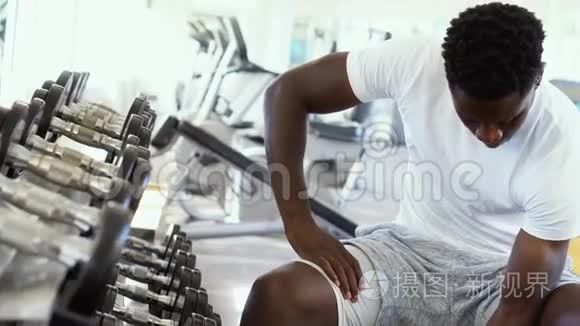年轻的非裔美国人坐着举起一个哑铃，靠近健身房的架子。 男性体重训练者做