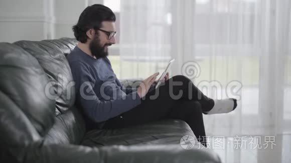 戴眼镜的白种人的侧视图，使用平板电脑和微笑。 年轻人坐在舒适的沙发上