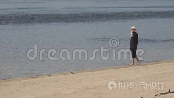 戴着太阳镜戴着帽子的人正慢慢地沿着海滩上的沙子沿着水走，在打电话