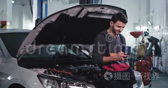 一个穿制服的工人微笑着在汽车修理厂修理汽车时看了看手机