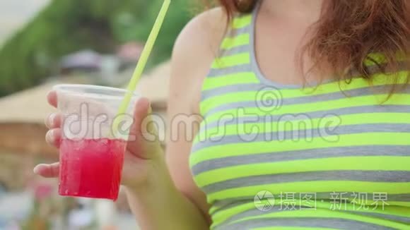 在阳光明媚的日子里，年轻的女人手里拿着红色的鸡尾酒和冰。 近距离的女人喝草莓鸡尾酒