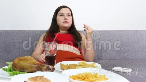 丰满的女人坐在桌子旁吃高热量的垃圾食品。 看电视。