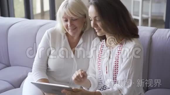 年轻的女人和她的母亲坐在沙发上，在平板电脑上展示她的照片。 女人微笑着。 友好家庭