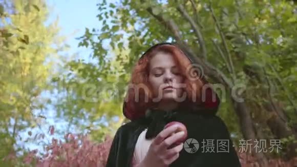 一个穿着黑色长袍的红发白种人女人的特写镜头看着她手中的苹果。 仙女站着