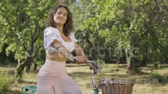 可爱的卷发年轻女子站在花园里或公园里，骑着自行车，看着镜头。农村生活